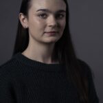 Professional headshot of young woman in dark sweater against a grey background