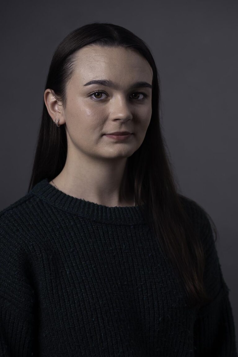 Professional headshot of young woman in dark sweater against a grey background